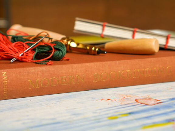 Book binding tools lay on top of a book in a rust colour, that reads "Modern Bookbinding".