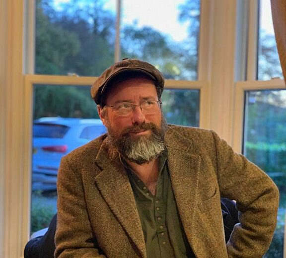 Image shows Ralph Bullivant sitting at a table with a knife and fork in front of him. They are wearing a brown jacket and hat, have a beard and glasses, and are sat in front of a window which looks out to cars and trees.