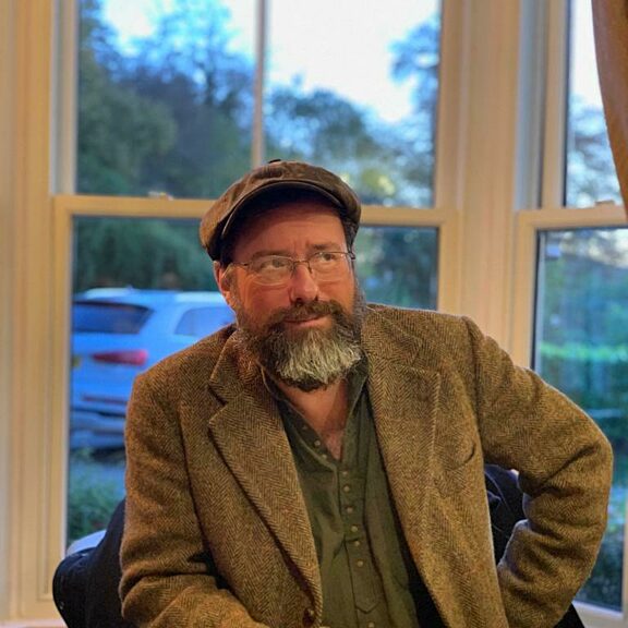 Image shows Ralph Bullivant sitting at a table with a knife and fork in front of him. They are wearing a brown jacket and hat, have a beard and glasses, and are sat in front of a window which looks out to cars and trees.