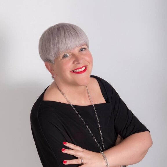 Image shows Sally-Anne smiling at the camera with her arms crossed. They are wearing a black dress, a long necklace and red lipstick. They have short white/grey hair and are standing in front of a white background.