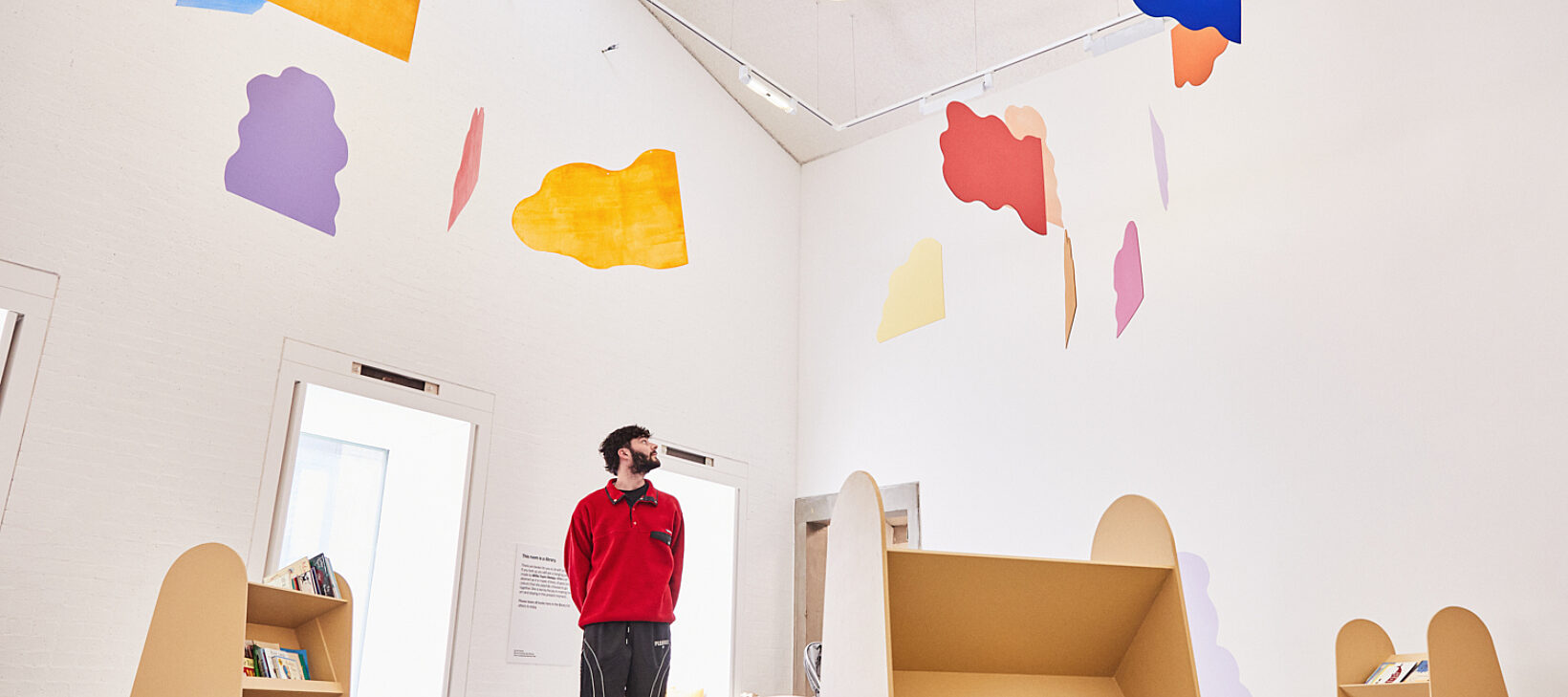 A colourful mobile made out of cloud shapes suspended in a white-walled room. A man in a red fleece looks at it.