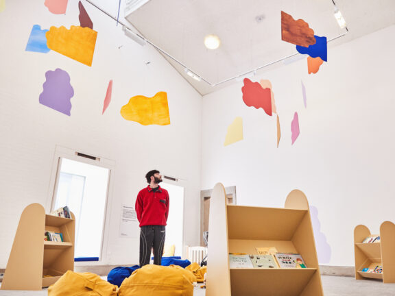 A colourful mobile made out of cloud shapes suspended in a white-walled room. A man in a red fleece looks at it.