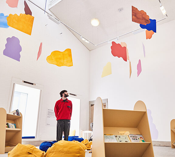 A colourful mobile made out of cloud shapes suspended in a white-walled room. A man in a red fleece looks at it.
