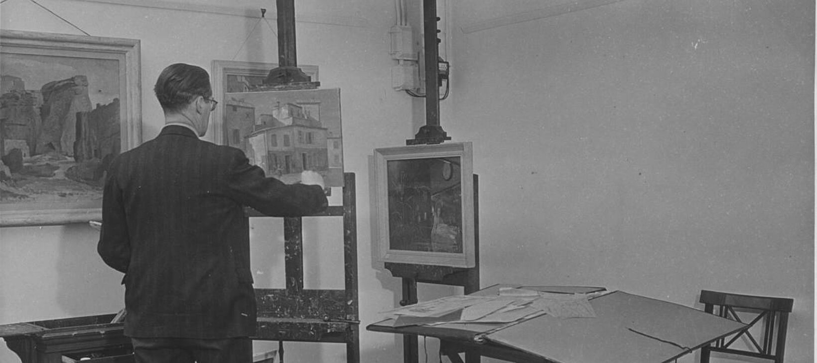 An artist stands with their back to the camera facing an easel. There are other paintings in the background and a table and chair in the room.
