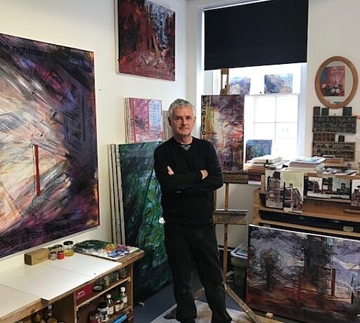 Person dressed in all black stands with their arms crossed in the middle of an art studio, surrounded by paintings on the wall.