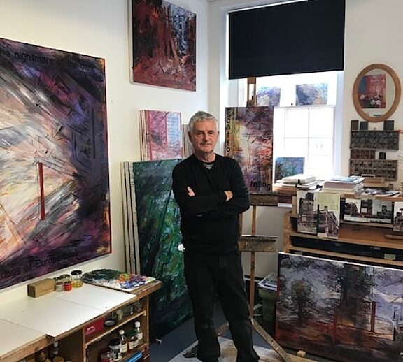 Person dressed in all black stands with their arms crossed in the middle of an art studio, surrounded by paintings on the wall.