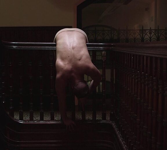 A person is bended over a dark wood bannister in a dark hallway.