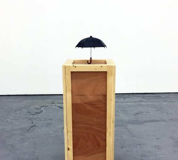 A wooden block stands against a white wall and grey background, with a small black umbrella balanced on top