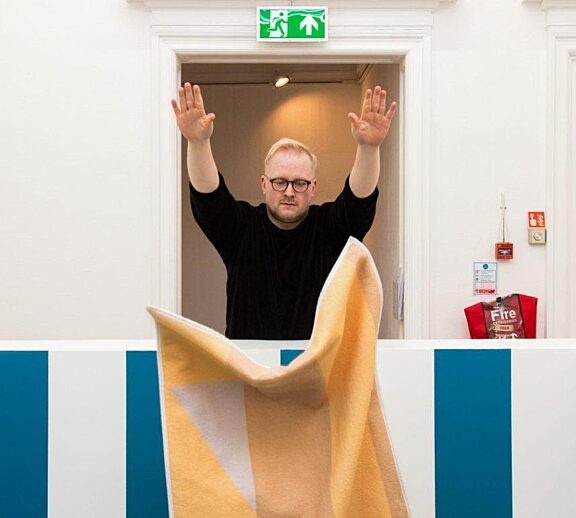 A person dressed in a black jumper throws a yellow piece of fabric over a blue and white wall. They are stood in front of a doorway.