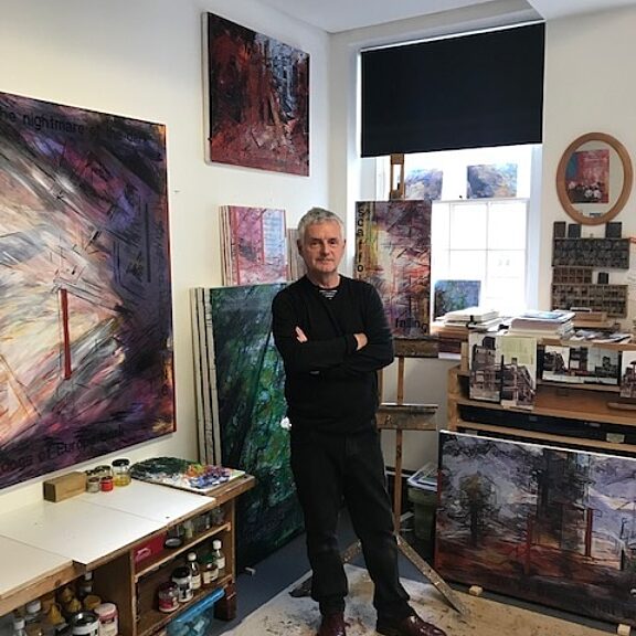 Person dressed in all black stands with their arms crossed in the middle of an art studio, surrounded by paintings on the wall.