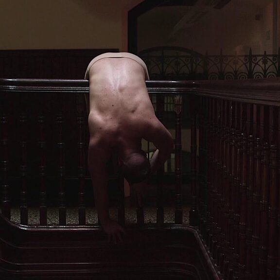 A person is bended over a dark wood bannister in a dark hallway.
