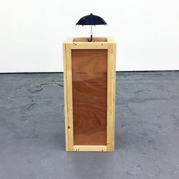 A wooden block stands against a white wall and grey background, with a small black umbrella balanced on top