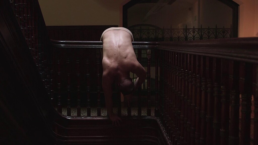 A person is bended over a dark wood bannister in a dark hallway.