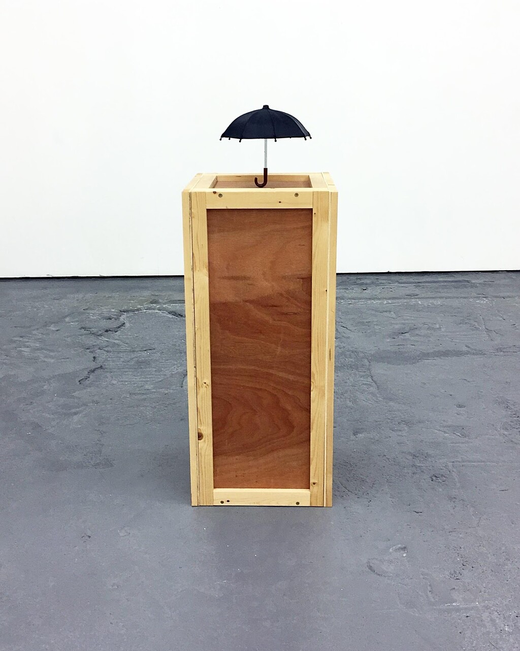 A wooden block stands against a white wall and grey background, with a small black umbrella balanced on top