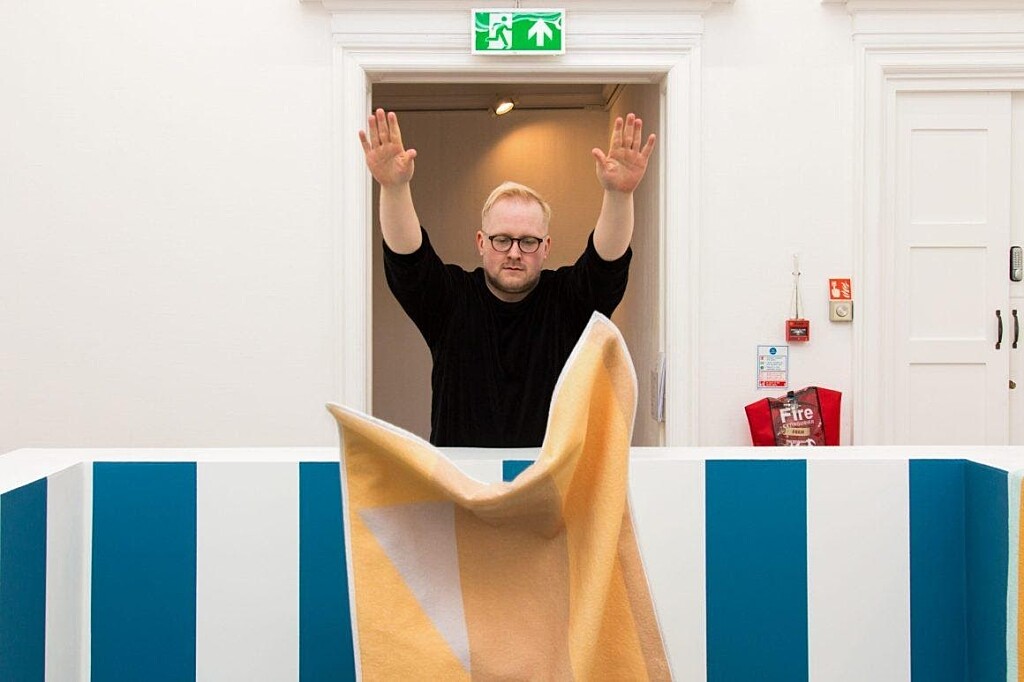A person dressed in a black jumper throws a yellow piece of fabric over a blue and white wall. They are stood in front of a doorway.