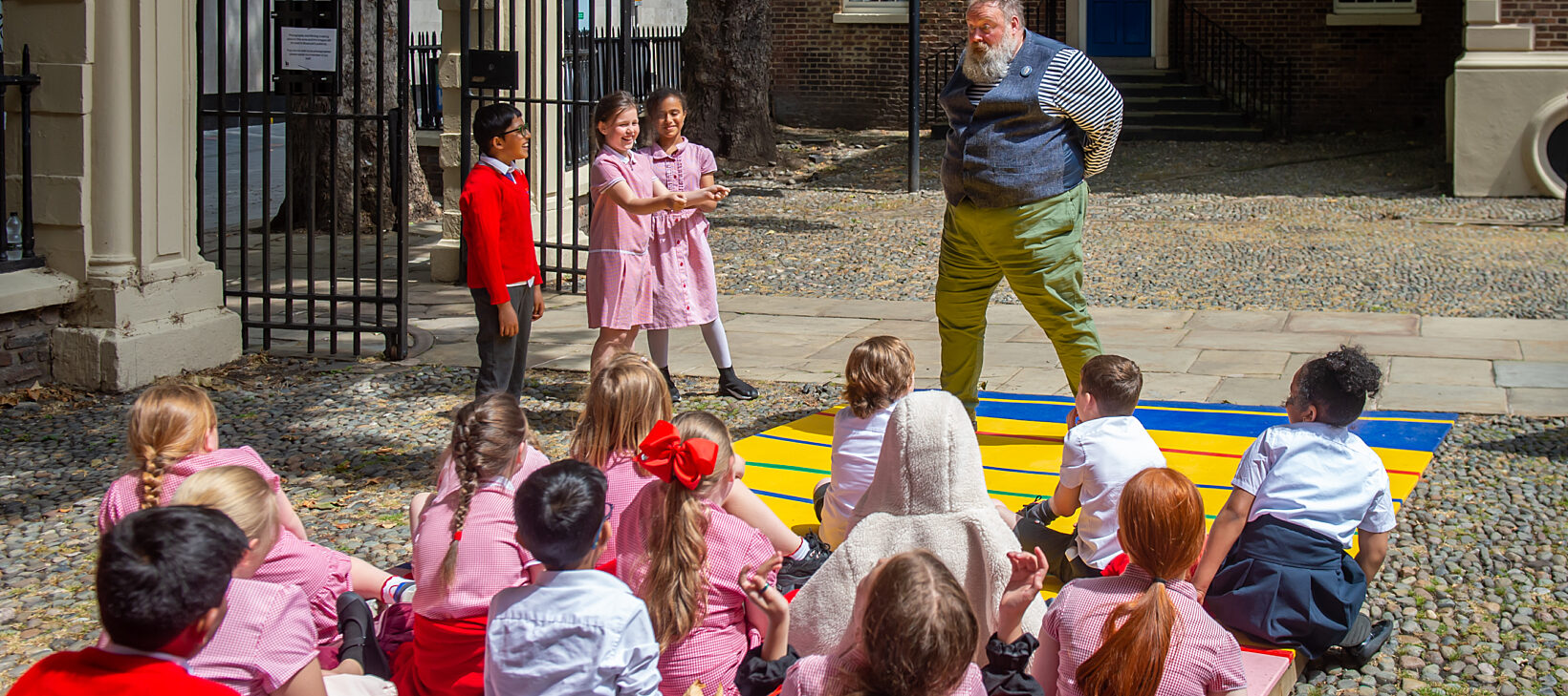 A group of primary school children gather in front of a performer, who is wearing a waist jacket, outside of the Bluecoat building.