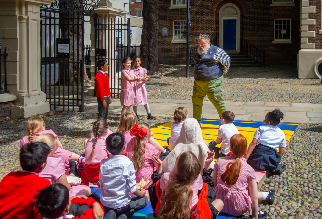 A group of primary school children gather in front of a performer, who is wearing a waist jacket, outside of the Bluecoat building.