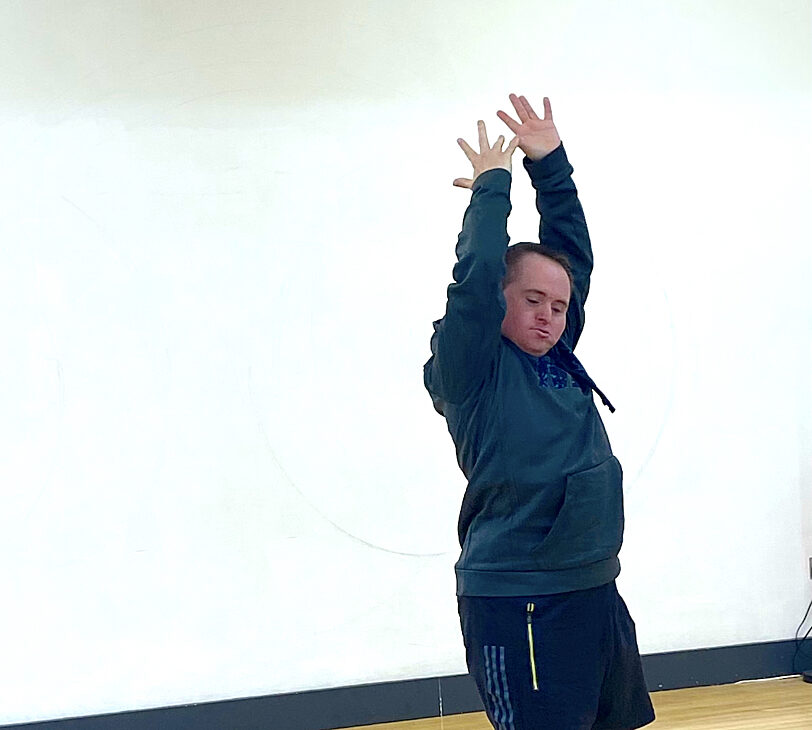 Image shows Blue Room artist Tony Carroll in a dance studio. He is wearing a blue jumper and black shorts and has his arms raised above his head with his eyes closed.