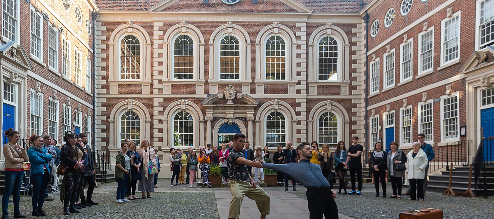 Two dancers perform in Bluecoat's courtyard with a packed audience behind them. In the background you can see the Bluecoat building.