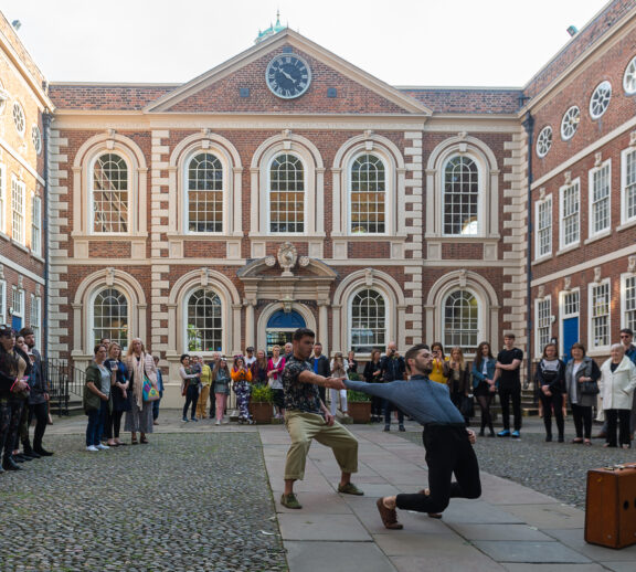 Two dancers perform in Bluecoat's courtyard with a packed audience behind them. In the background you can see the Bluecoat building.