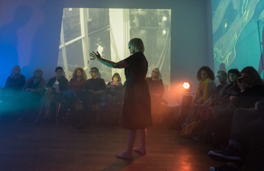 The room is dark, with film projection on the wall. There are two rows of people, both facing into a dance floor where a performer is standing in dimly lit spot lights, raises their hand.