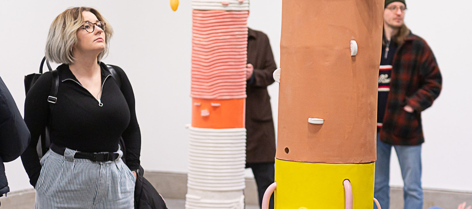 A woman wearing a black top and grey trousers looks up at a brown, black and yellow sculpture. There are two people in the background who are also looking the artwork.