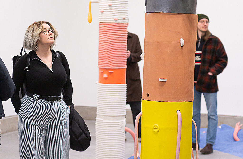 A woman wearing a black top and grey trousers looks up at a brown, black and yellow sculpture. There are two people in the background who are also looking the artwork.