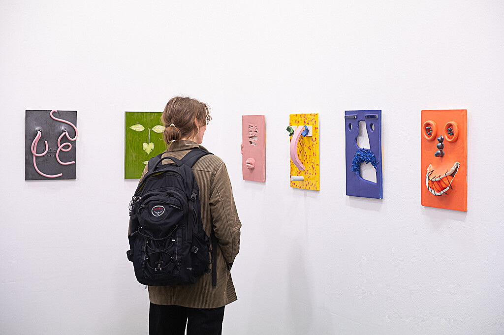 A photograph showing Jonathan Baldock's exhibition Facecrime. The image shows a visitor looking at a selection of works hanging on a wall. Each piece is a small vibrantly-coloured ceramic depiction of a face.
