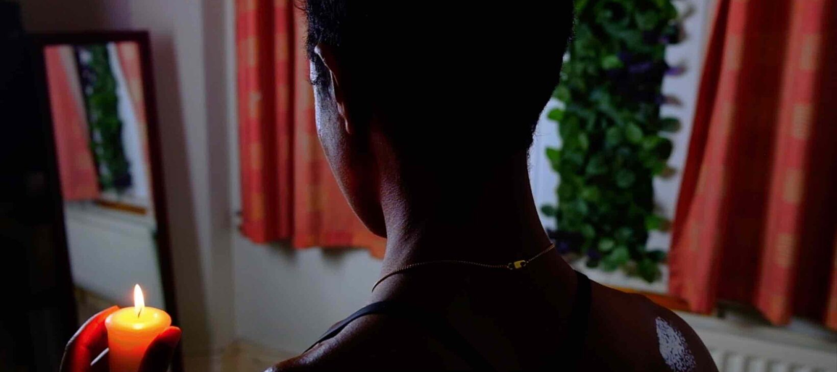 A film still showing a black person facing away from the camera and holding a lit candle. The subtitles read, "we are living through an upheaval."