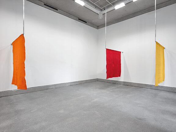 An installation shot of Undo Things Done, featuring three hanging quilts, one orange, one red, and one yellow.