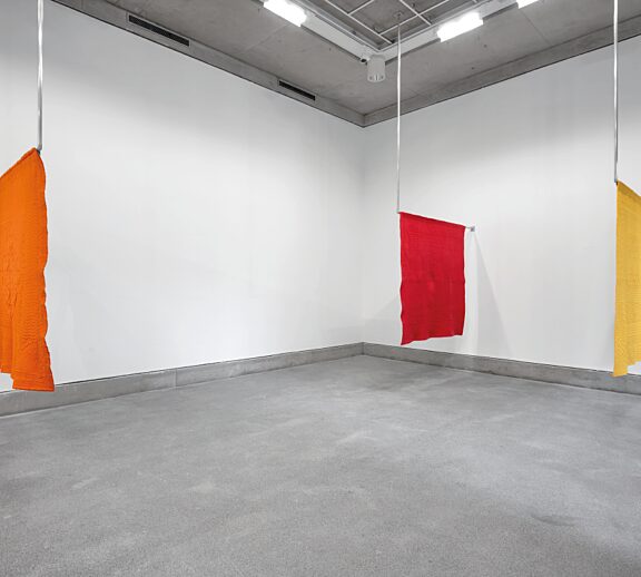 An installation shot of Undo Things Done, featuring three hanging quilts, one orange, one red, and one yellow.