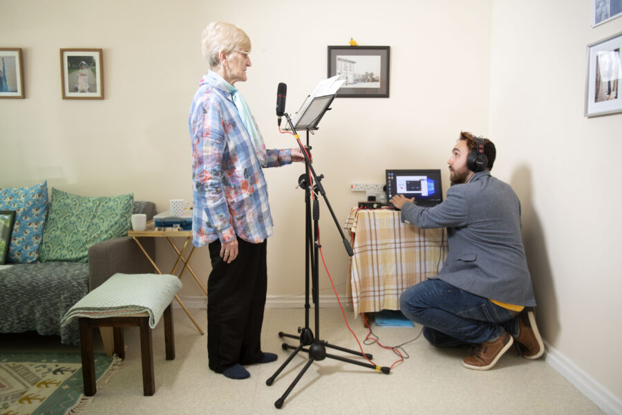 Image shows an older white woman, with a script, speaking into a microphone, while a younger white man wearing headphones with a laptop watches her.