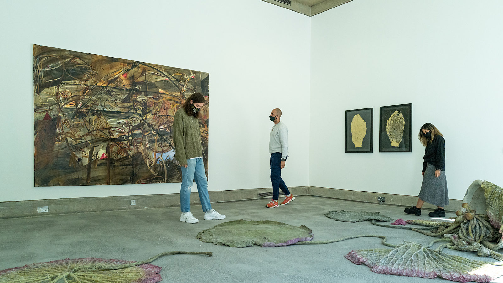 Three people are walking around a gallery space with paintings on the wall and sculptures on the floor.