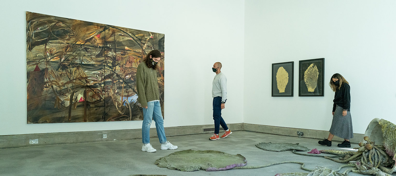 Three people are walking around a gallery space with paintings on the wall and sculptures on the floor.