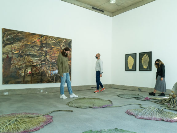 Three people are walking around a gallery space with paintings on the wall and sculptures on the floor.