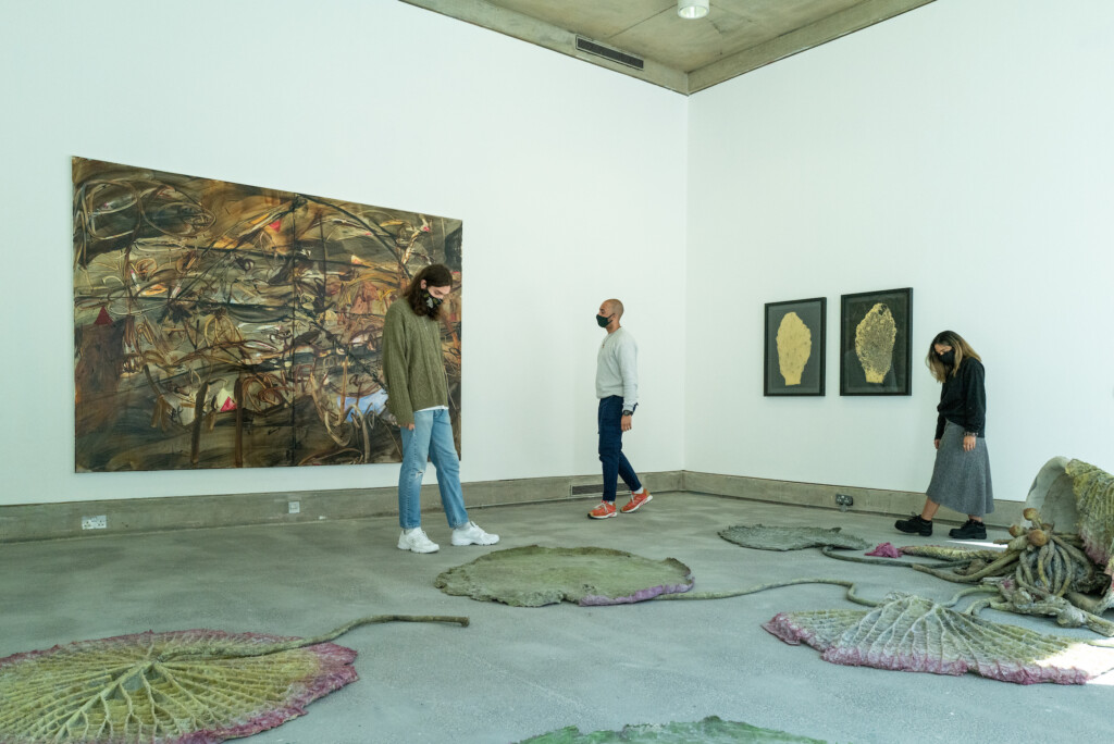 Three people are walking around a gallery space with paintings on the wall and sculptures on the floor.