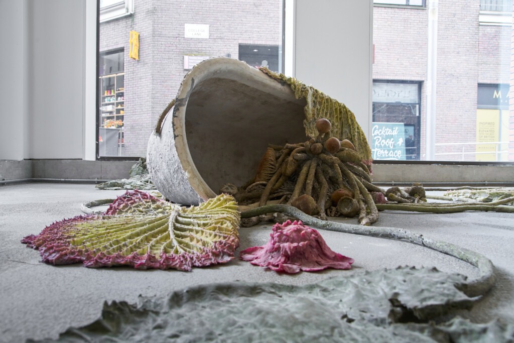 A close-up of a large sculptural work. The piece depicts giant lily pads spilling out of a bath and onto the gallery floor.