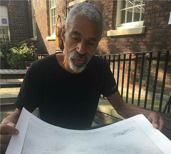 Tony Phillips sits at a table in Bluecoat Garden, he is holding a large piece of white paper with grey etchings on it. He is wearing a black tshirt and is looking down at the artwork.