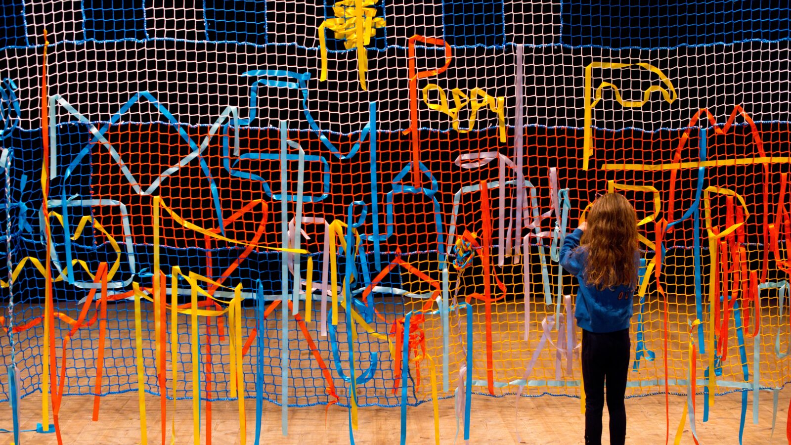 Image shows a lots of coloured fabric entangled in a large mesh wall. There is a young child with long brown hair with their back to the camera playing with the fabric.
