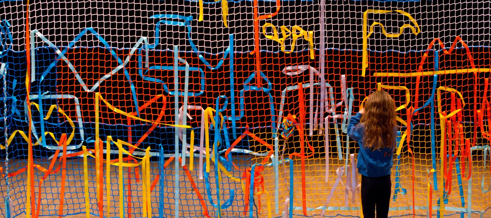 Image shows a lots of coloured fabric entangled in a large mesh wall. There is a young child with long brown hair with their back to the camera playing with the fabric.