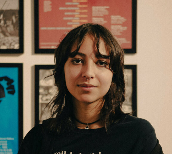 Image is a portrait photo of Shanti. She is wearing a black tshirt that has the text 'well behaved women barely make history.', a black necklace and has black short hair.