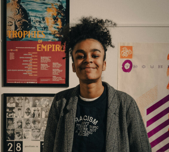 Image is a portrait of Tyrah stood in the gallery. They are looking straight towards the camera and are wearing a black tshirt with and grey jacket. They have black hair.