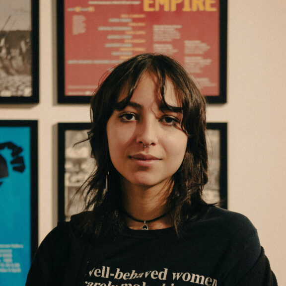 Image is a portrait photo of Shanti. She is wearing a black tshirt that has the text 'well behaved women barely make history.', a black necklace and has black short hair.