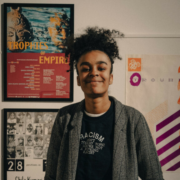 Image is a portrait of Tyrah stood in the gallery. They are looking straight towards the camera and are wearing a black tshirt with and grey jacket. They have black hair.