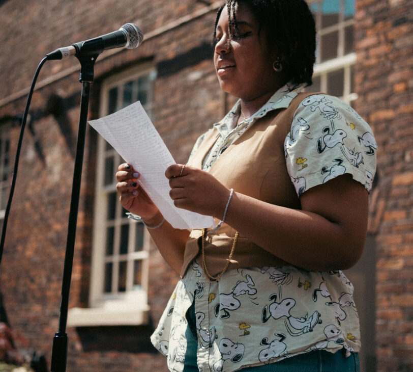 Image shows Nia stood behind a microphone holding a piece of paper, performing.