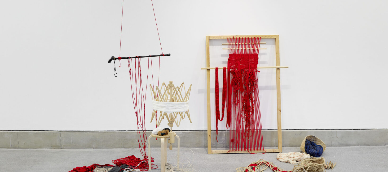 Image shows Raisa Kabir's art instillation. A wooden structure with red cloth, hanging down. To the left is a walking stick suspended by red cloth and ropes run along the floor.