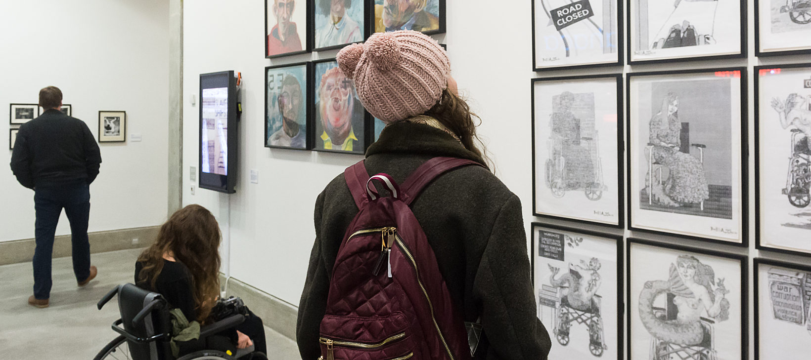 Two people, one in a wheel chair and another in a pink hat and purple, look toward a series of black and white images on the wall.