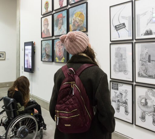 Two people, one in a wheel chair and another in a pink hat and purple, look toward a series of black and white images on the wall.