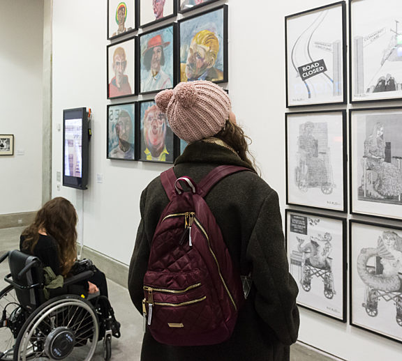 Two people, one in a wheel chair and another in a pink hat and purple, look toward a series of black and white images on the wall.