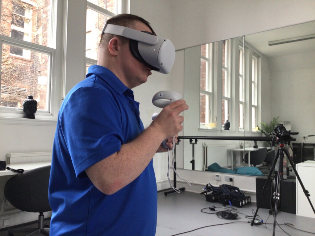 Image shows dance artist Tony Carroll wearing a VR headset and holding a VR controller in a white studio space.